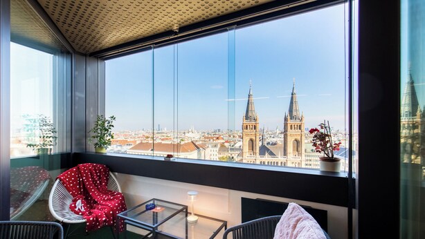 A balcony with a view of a cityscape, including two prominent towers. There are two chairs, one with a red polka-dotted blanket, and a small table with a lamp. - Roof-top panorama suite with distant views over Vienna (Wien)