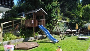 Une structure de jeu en bois comprenant un toboggan, une balançoire et des tables de pique-nique, installée dans une cour arrière verdoyante.