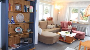 Un salon chaleureux avec une bibliothèque en bois, un canapé beige, un fauteuil rose et une petite table sur laquelle se trouvent une bouteille et des verres.