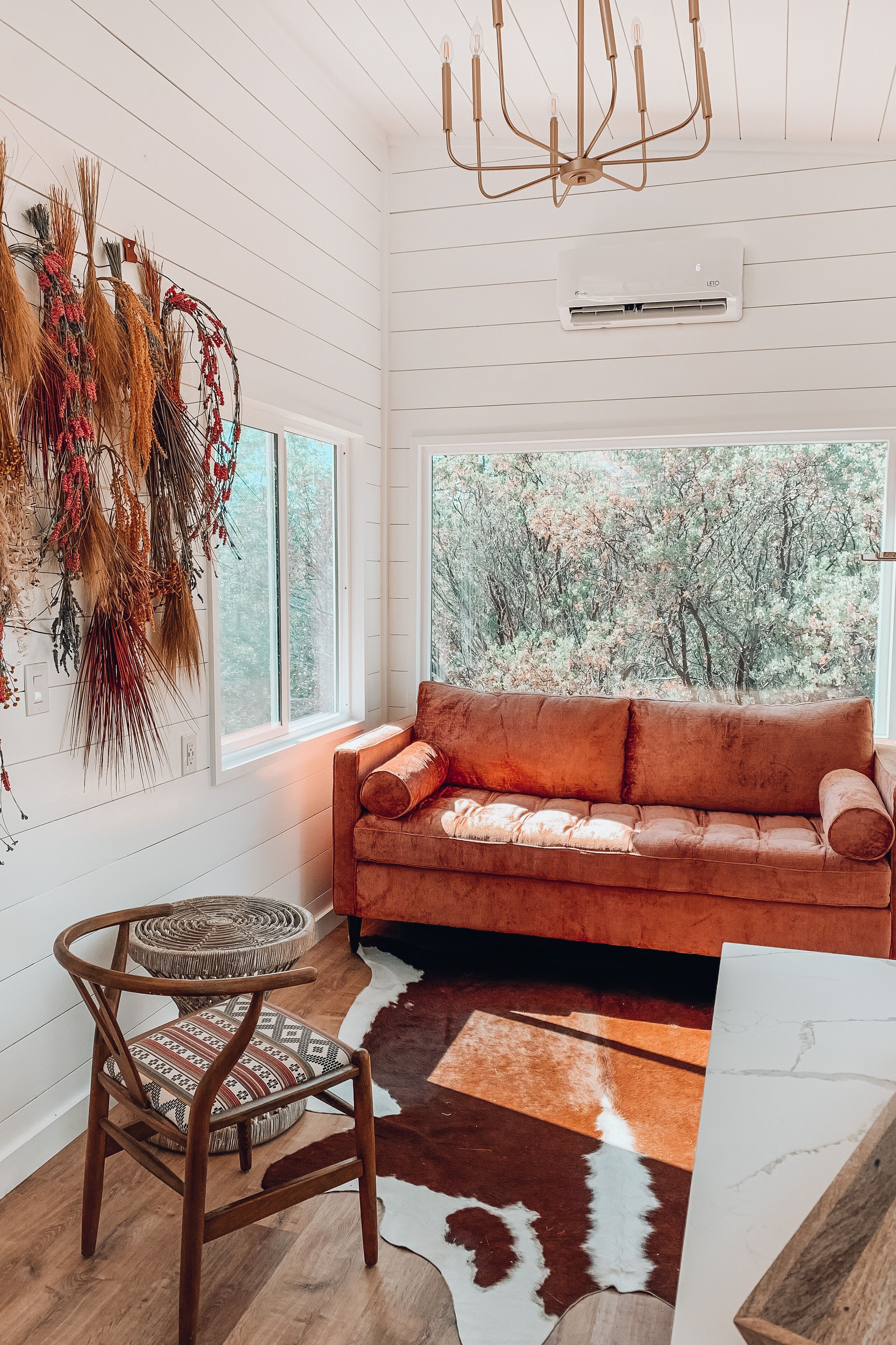 A cozy room with a wooden floor, a brown sofa, a patterned chair, and a large window with a view of trees.