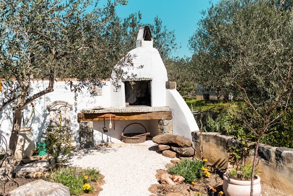 A traditional stone oven with a white dome roof, surrounded by olive trees and a gravel path.