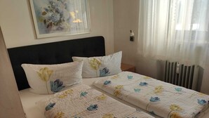 A hotel room with two beds, each with floral-patterned bedding and pillows. There is a framed artwork on the wall above the beds, a bedside table with a lamp, and a window with sheer curtains. - Apartment Englmari for 5 pers. with 60 sqm in St. Englmar (Sankt Englmar)