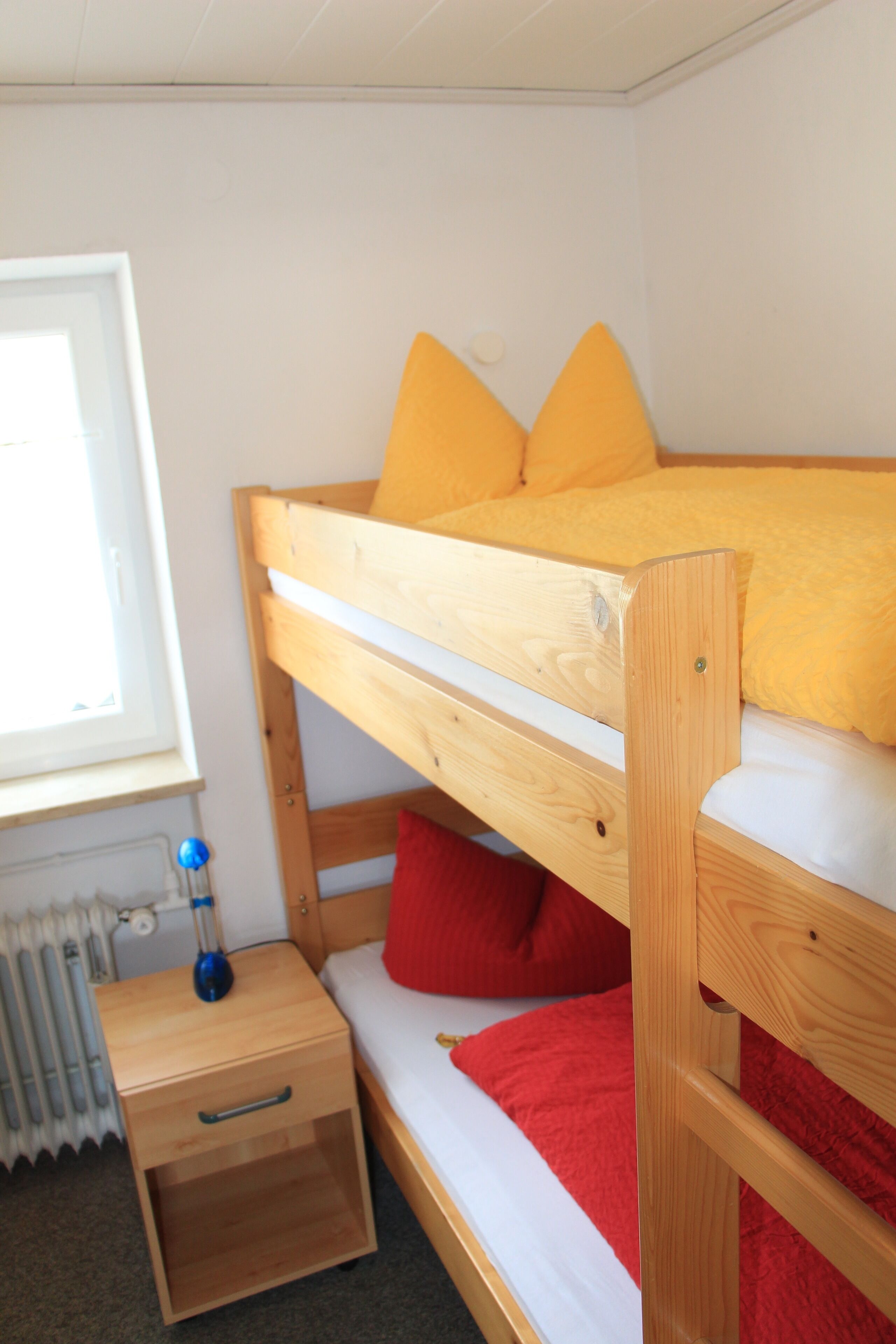 A bunk bed with a wooden frame, a small bedside table, and a window with a view of a radiator.