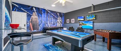 A pool table with a blue felt surface, a foosball table, and a bar area with a red bowl.