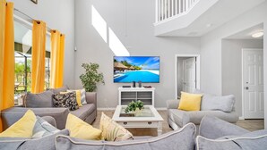 Een ruime woonkamer met een grote flatscreen-televisie, grijze en gele banken en een witte salontafel met een plant.