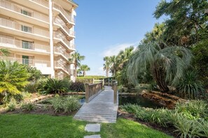 A wooden pathway leading to a building with multiple balconies, surrounded by lush greenery and a small pond.