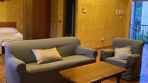 En hyggelig stue med en sofa, et træ sofabord og en lænestol. En dør fører til et andet rum, og der er en balkon, som kan ses gennem en åben dør.