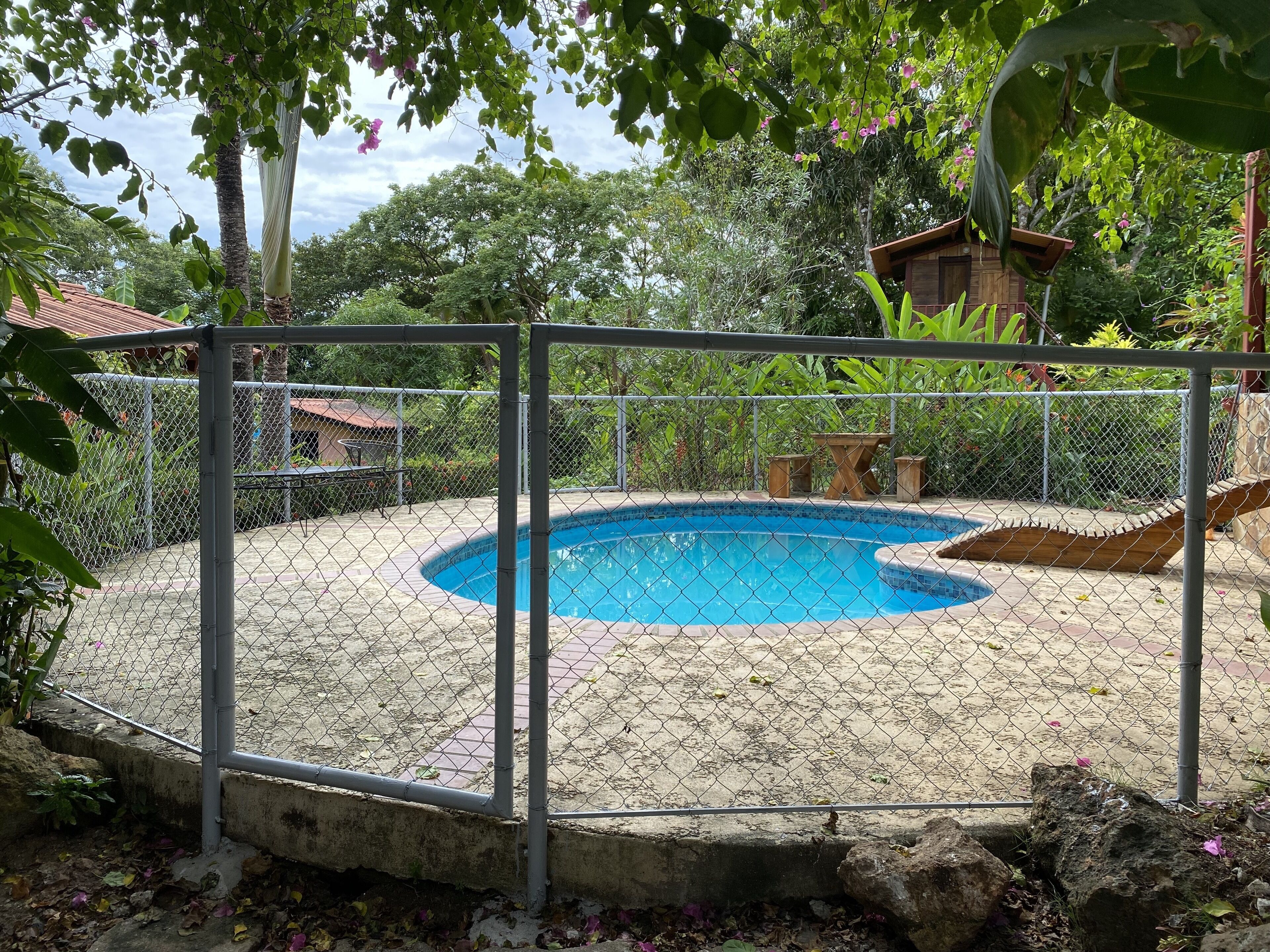 Una piscina envoltada per una tanca metàl·lica, amb un tramadol de fusta i una petita construcció de fusta al fons.