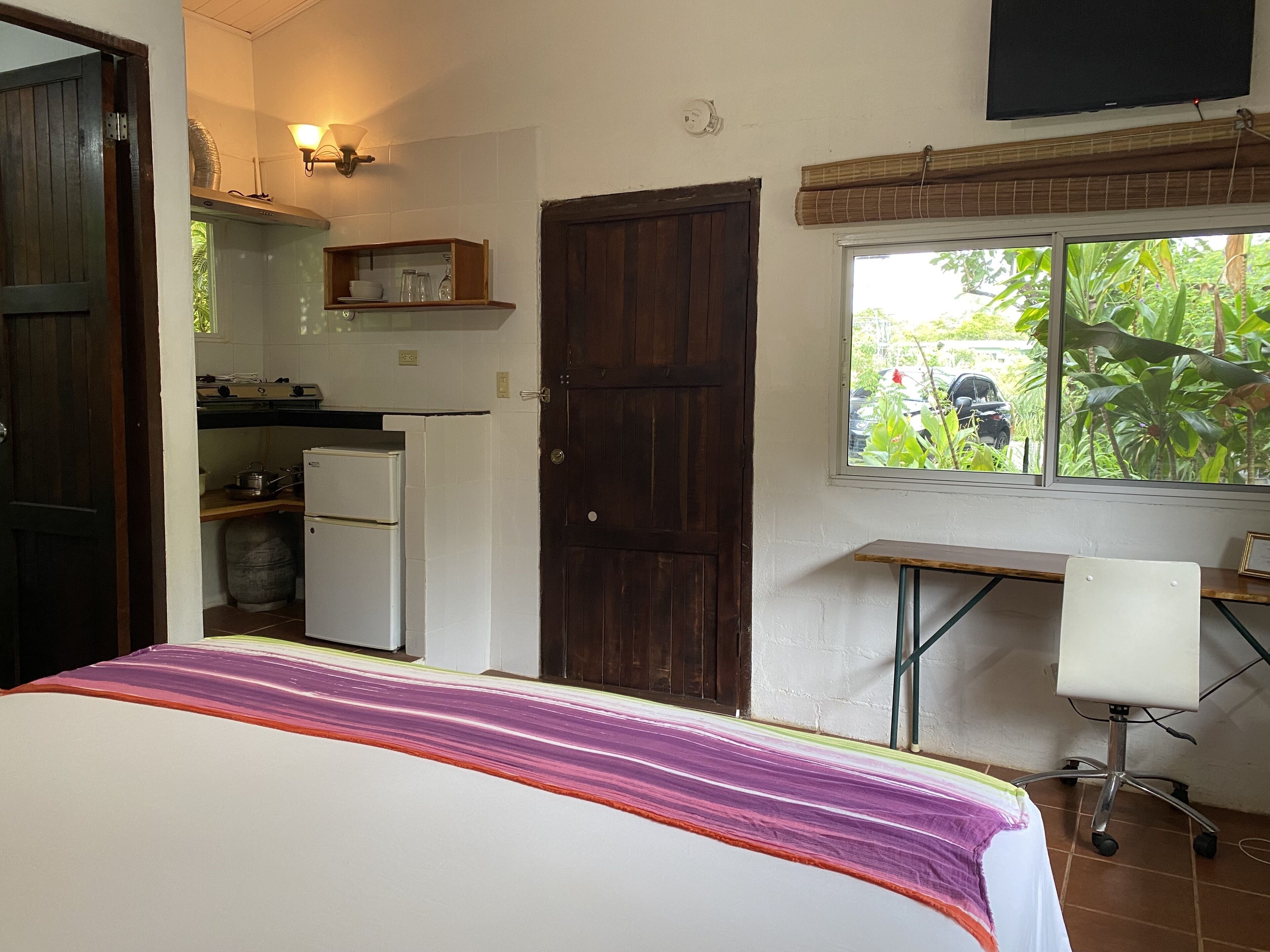 A bedroom with a bed, a desk, a chair, a window with a view of greenery, and a kitchen area with a refrigerator and stove.