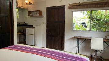 A bedroom with a bed, a desk, a chair, a window with a view of greenery, and a kitchen area with a refrigerator and stove.