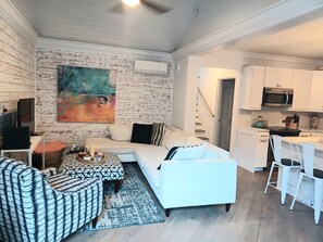 A modern living room with a white sofa, patterned ottoman, and a colorful abstract painting on the wall. A kitchen area with white cabinets and a microwave is visible in the background.