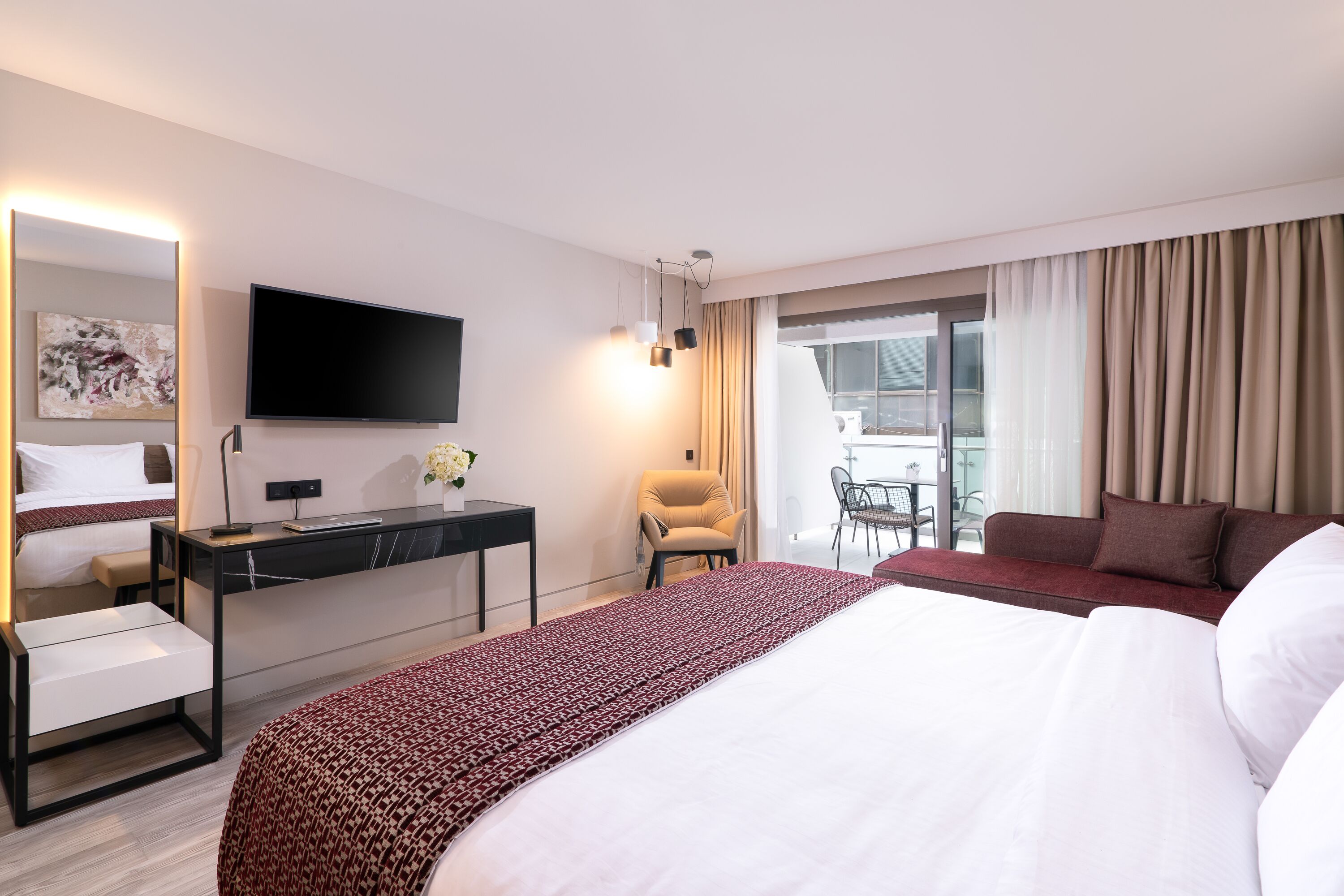 A hotel room with a large bed, a desk with a flat-screen TV, a chair, and a balcony with curtains.