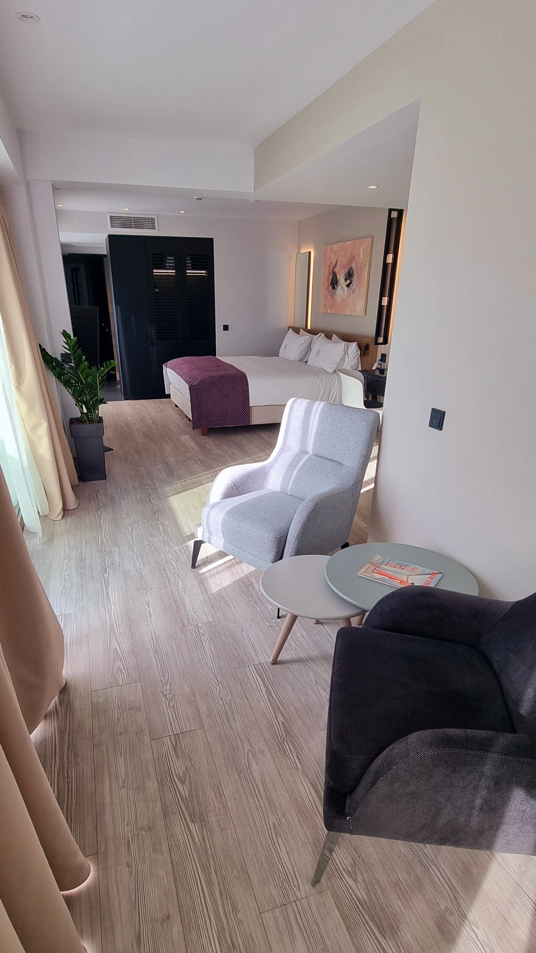 Una habitació d'hotel moderna amb una butaca gris, una taula rodona petita, un sofà, un llit amb roba de llit blanca, un mirall gran i una planta en test.