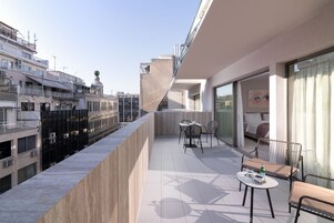 Un balcón con mesa y sillas, una cama con vistas y un paisaje urbano al fondo.