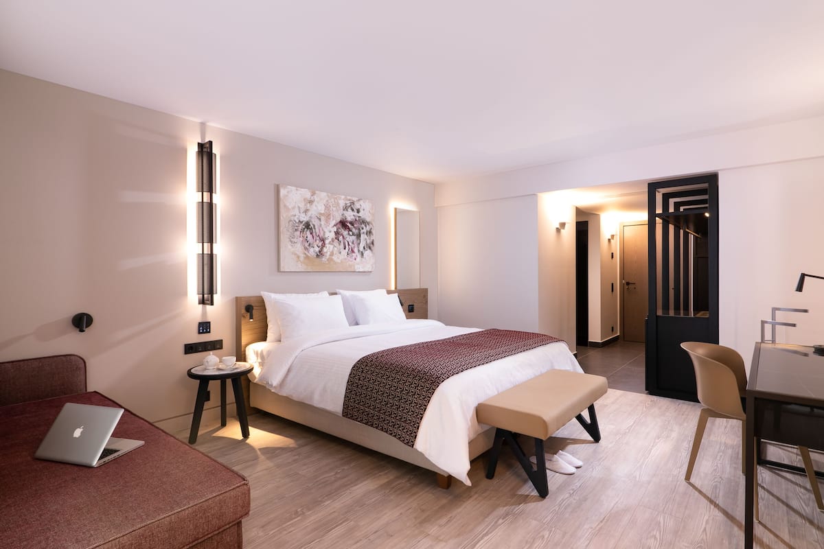 Moderni hotellihuone, jossa on sänky, yöpöytä, lamppu ja kannettava tietokone punaisella sohvalla.