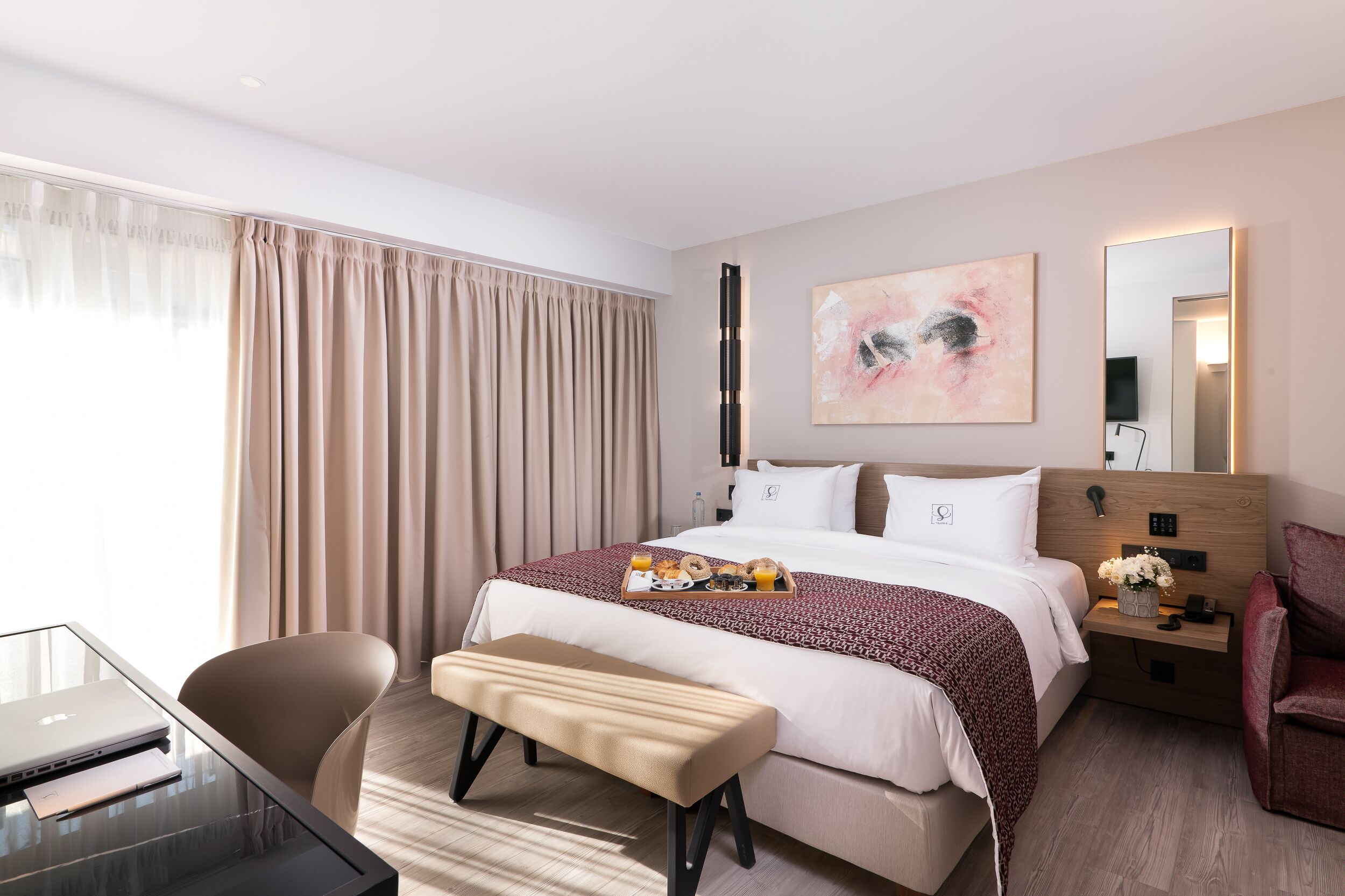 Una habitación de hotel con una cama, un escritorio con una computadora portátil, un banco, una mesita de noche con un jarrón de flores y una ventana grande con cortinas.