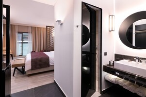 A modern hotel room with a large bed, a separate bathroom with a round mirror, and a seating area with a chair and table.