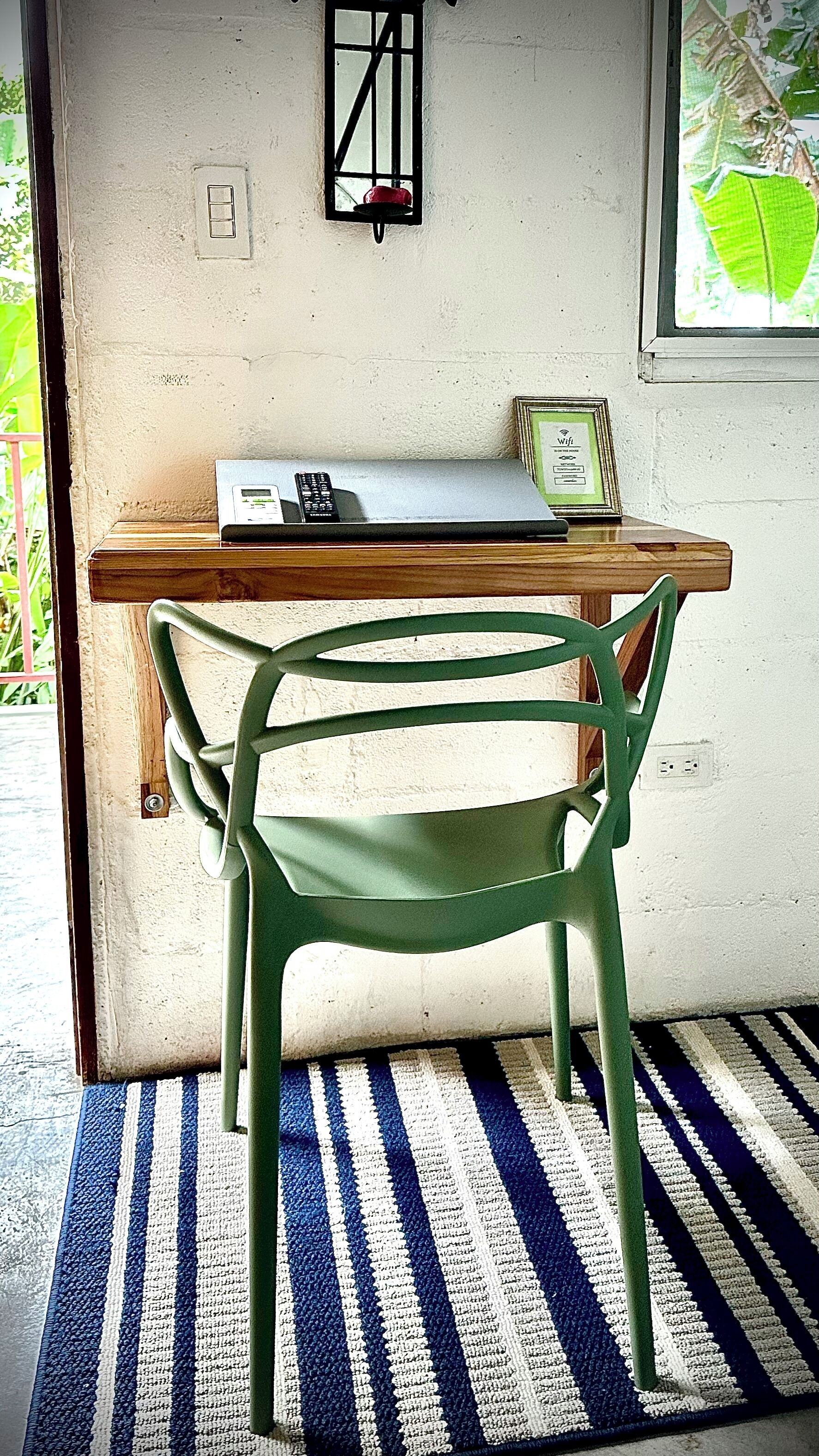 Una stanza con una scrivania in legno, una sedia verde, un tappeto a righe, una lampada a parete e una finestra con una pianta.