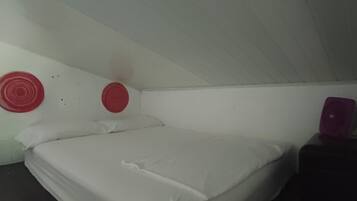 En säng för en person med vita sängkläder i ett rum med vita väggar och ett mörkt golv. Det finns två röda, runda väggdekorationer.