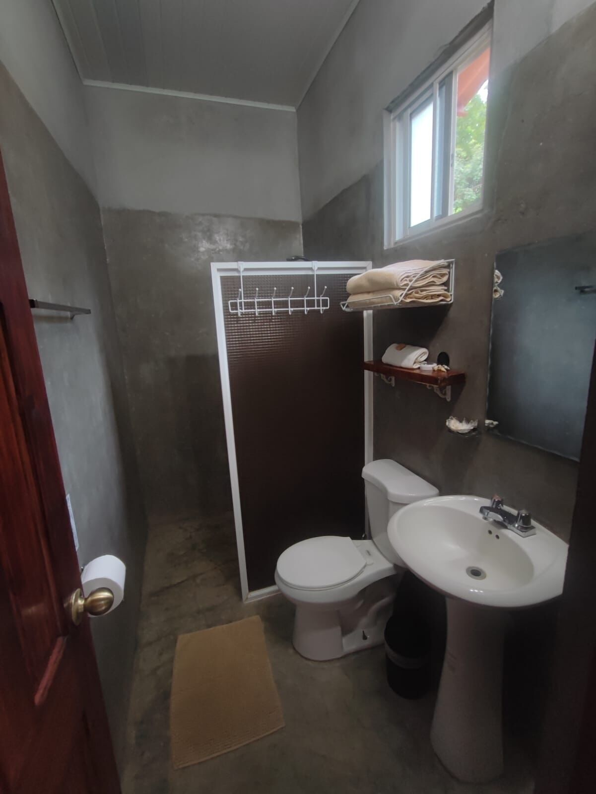 A small bathroom with a toilet, sink, and a shower stall. There is a towel rack above the sink and a window above the shower.