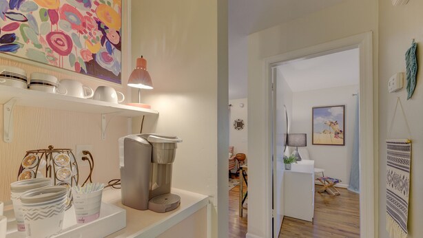 A kitchen with white cabinets, a coffee machine, and a hanging pot. There is a colorful painting on the wall and a door leading to a living room with a sofa and a table. - Comfy Cozy Convenient Condo (Columbia)