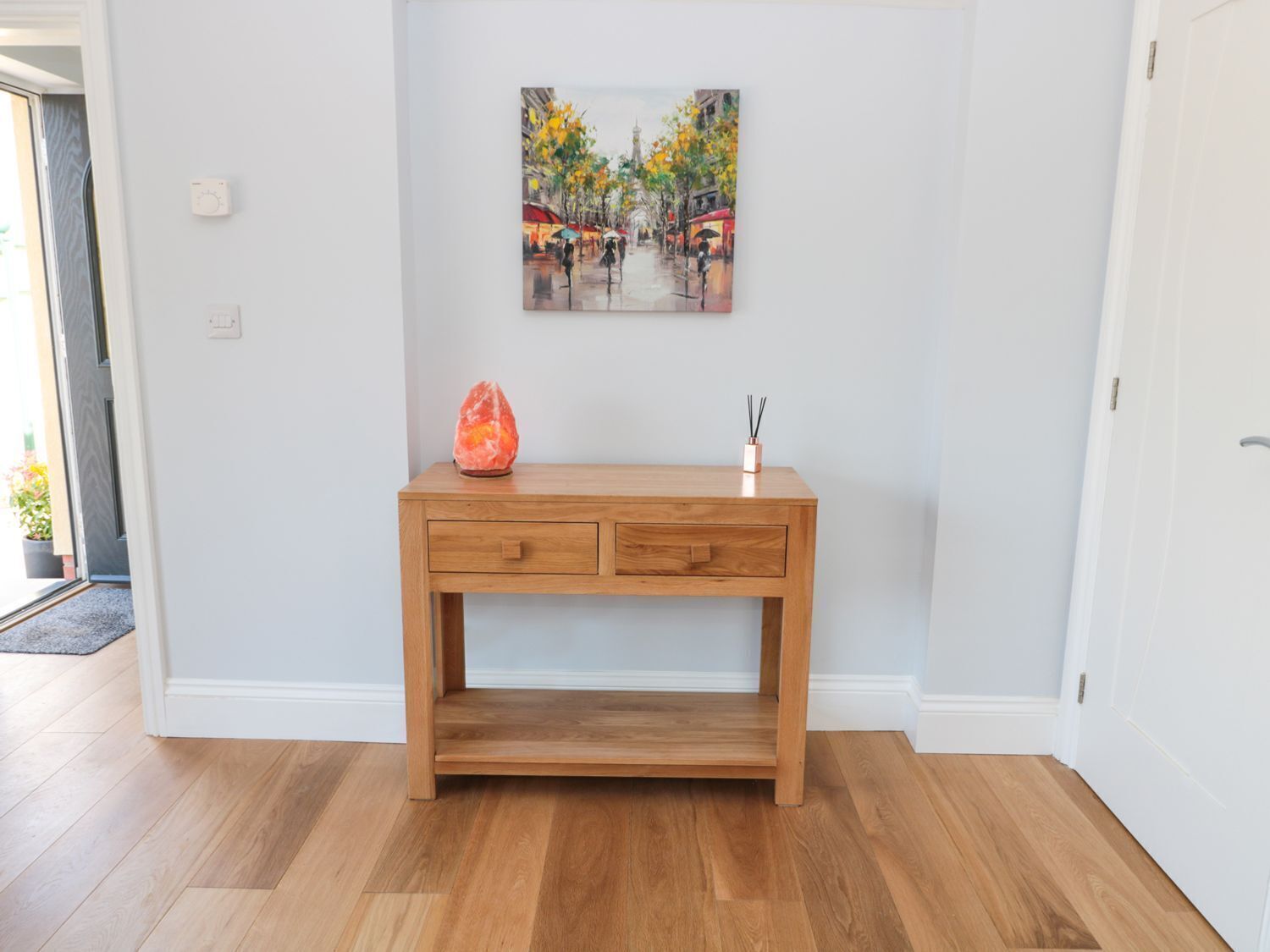 Une console en bois avec des tiroirs, une lampe de sel et un petit vase contenant des roseaux, le tout dans une pièce aux murs blancs et au plancher en bois massif.