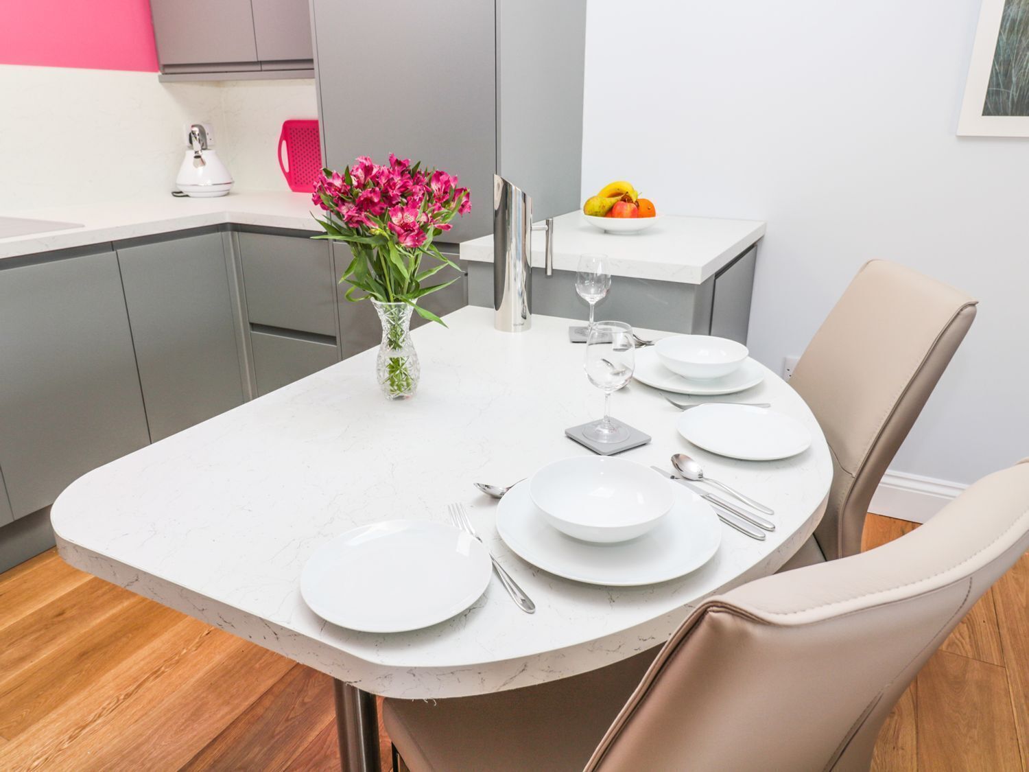 A modern kitchen with a dining table set for two, featuring white plates, silverware, and a vase of flowers.