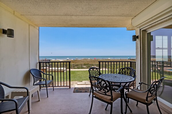 A balcony with a view of the beach, featuring a round table and chairs, a bench, and a railing.