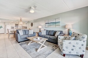 Ein modernes Wohnzimmer mit zwei Sofas, einem Couchtisch und einem gemusterten Sessel. Im Hintergrund ist eine Küche zu sehen.