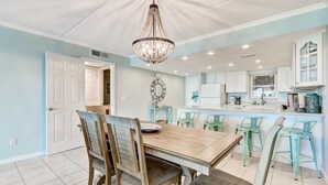 Un comedor con mesa y sillas de madera, una araña y una cocina con armarios blancos y un ventanal.