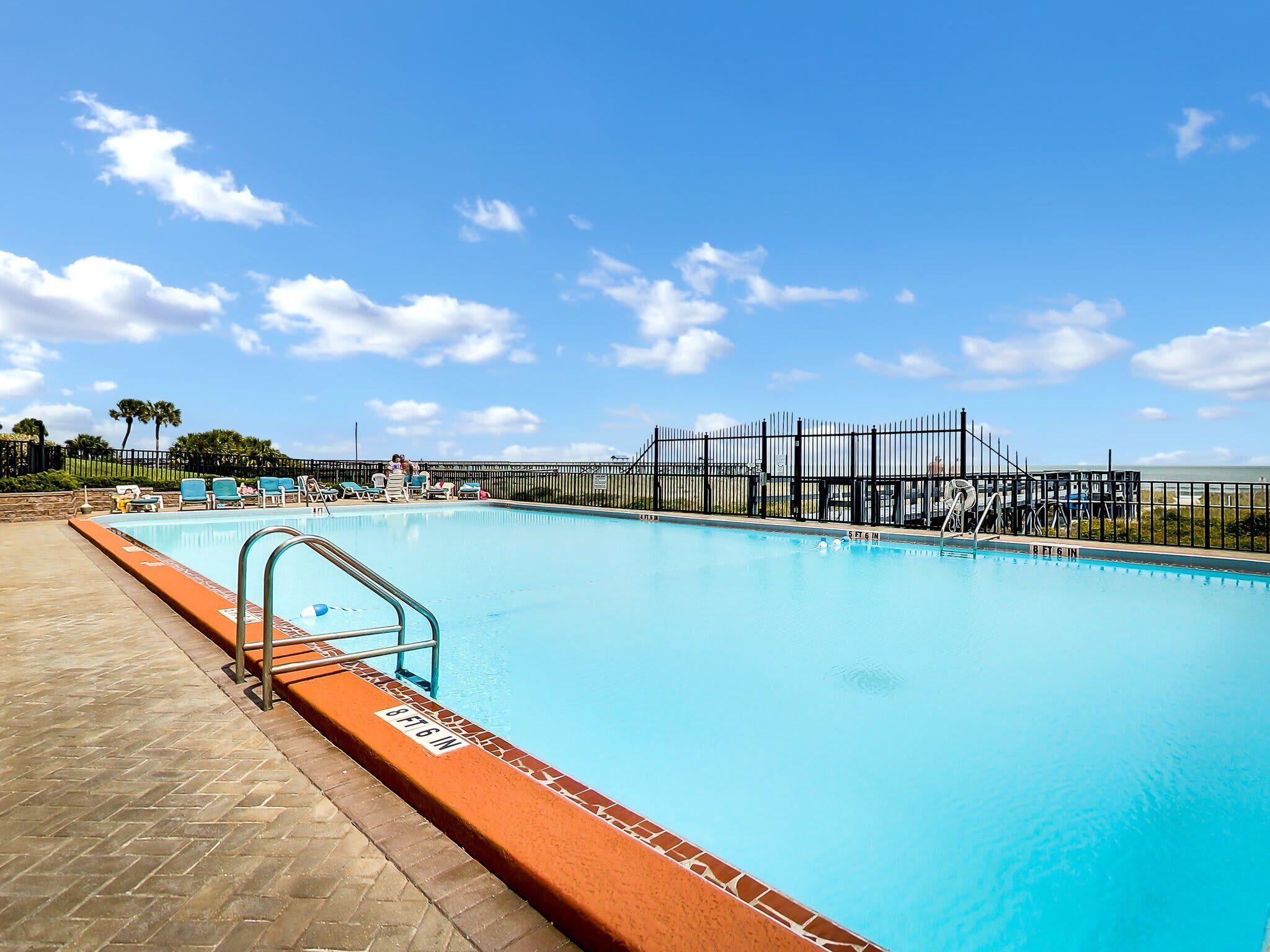 Une piscine avec un ciel bleu dégagé, une zone délimitée par une clôture et un chemin pavé.