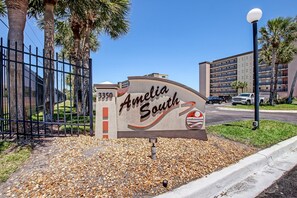 Schild für 'Amelia South' mit dem Logo einer Eigentumswohnung, umgeben von einem Zaun, Palmen und einem Parkplatz.