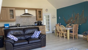 A modern living room with a leather sofa, a dining area with wooden chairs, and a kitchen with wooden cabinets and a tiled backsplash. - Awel Deg, Newport (Newport)