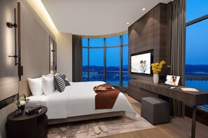 A modern hotel room with a large bed, a desk, a TV, and a view of the city at dusk. - Southbay Hotel Residence Zhuhai (Zhuhai)