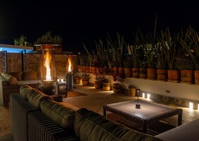 Un área de patio al aire libre con mobiliario, un elemento decorativo de fuego y plantas en macetas.