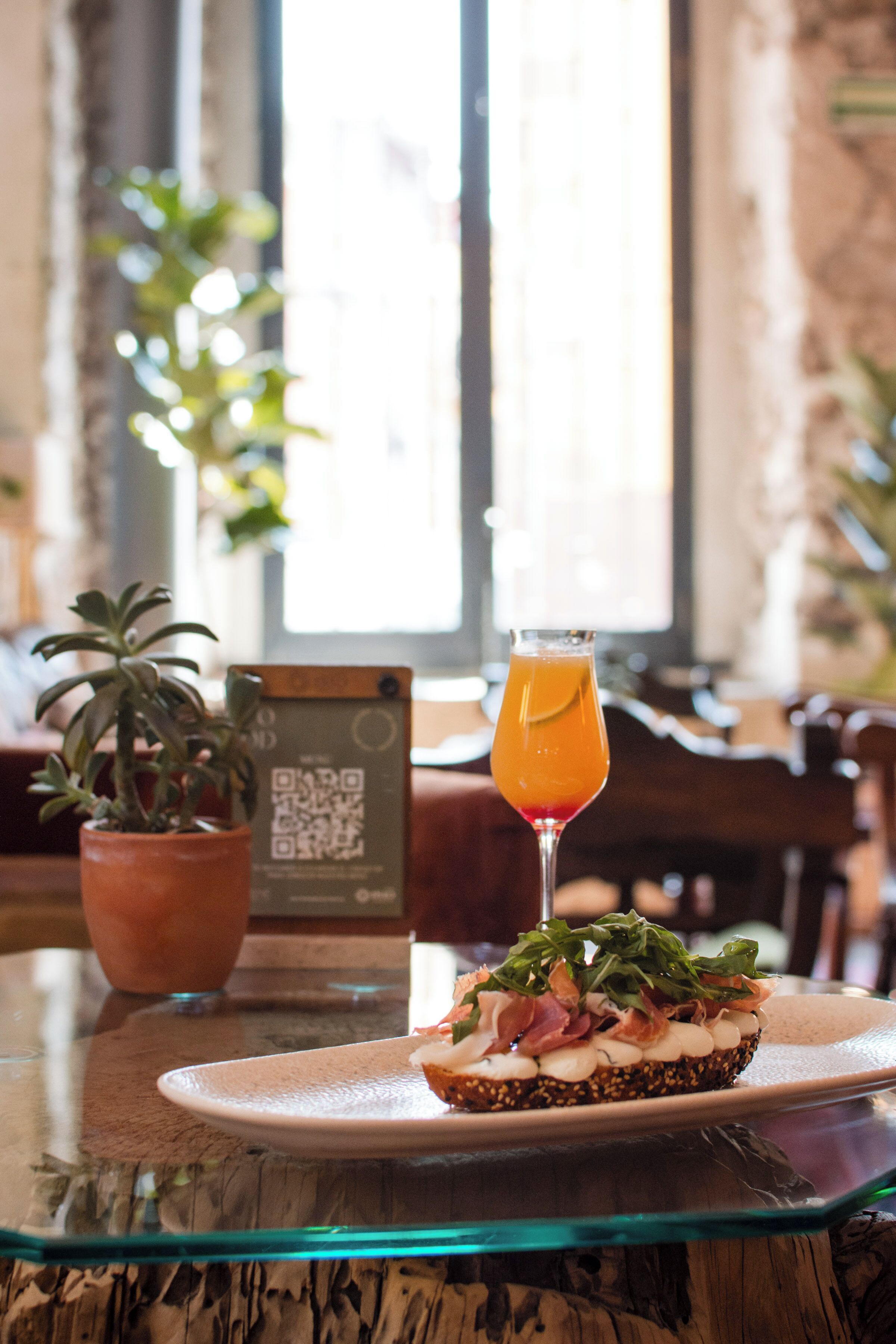 Ein Tisch mit einem Glas Orangensaft, einem Teller mit einem Sandwich und einem Karton mit einem QR-Code.