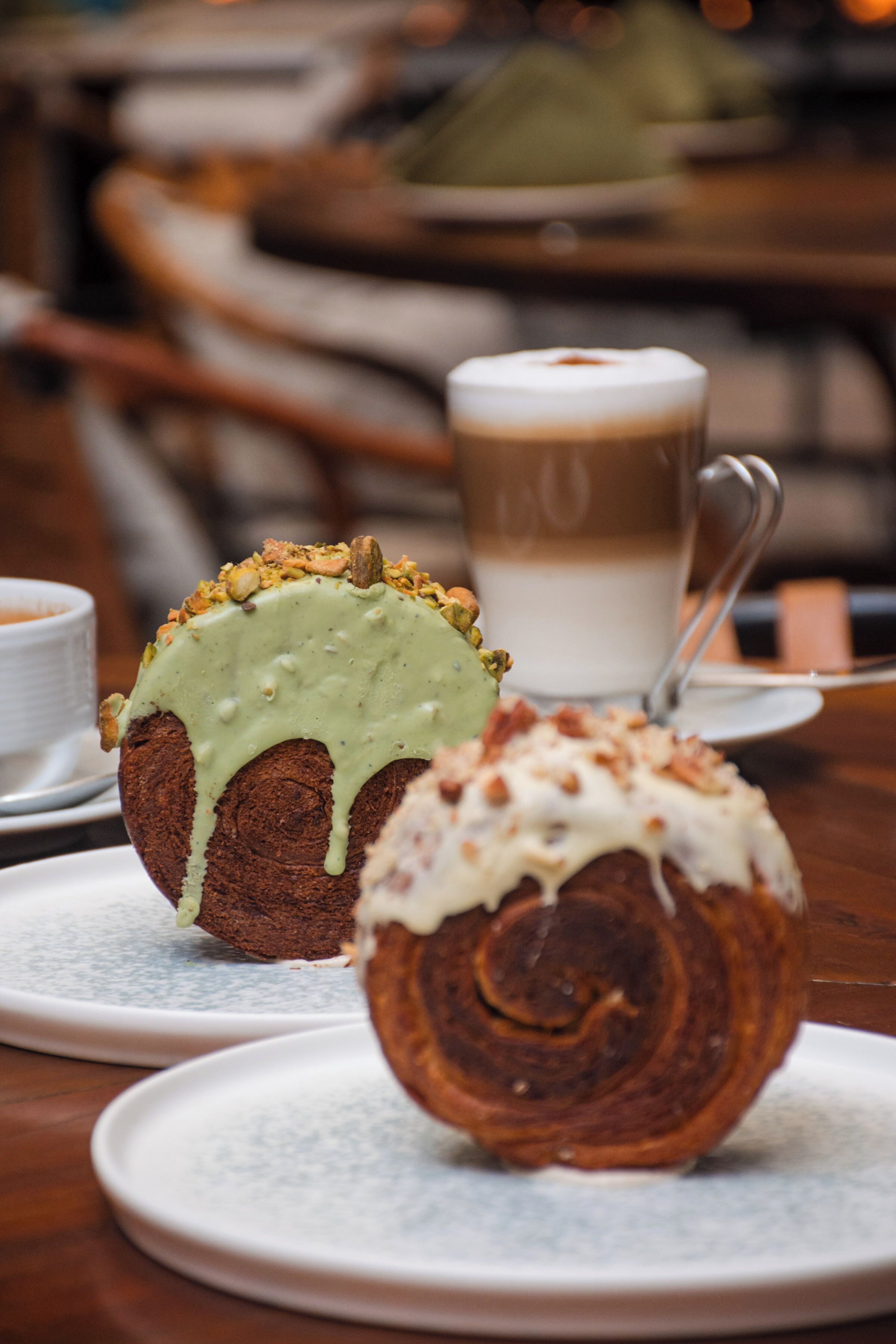 Une pâtisserie recouverte d’un glaçage vert et de noix, présentée sur une assiette, une autre pâtisserie enroulée avec un glaçage blanc, et une tasse de café en arrière-plan.