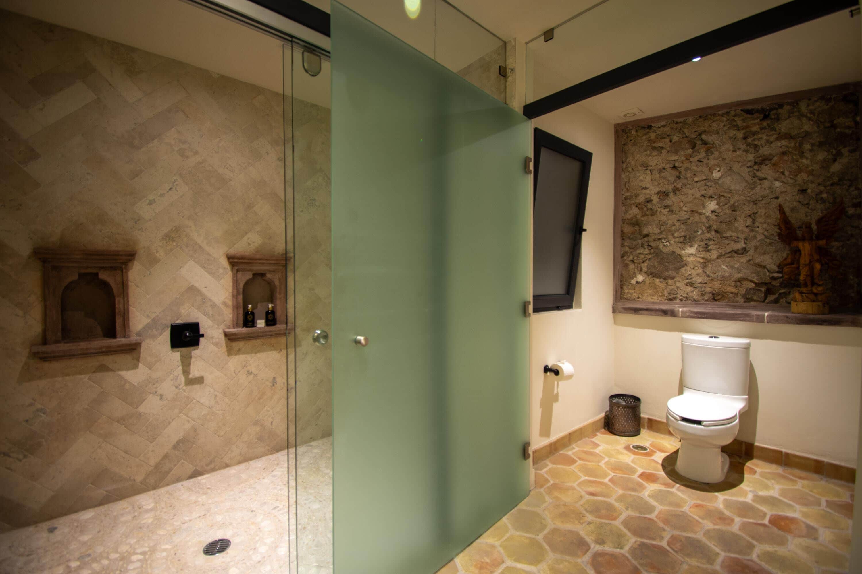 Un bagno con pavimento in pietra a mosaico a spina di pesce, cabina doccia in vetro, un water e una parete in pietra con un quadro appeso.