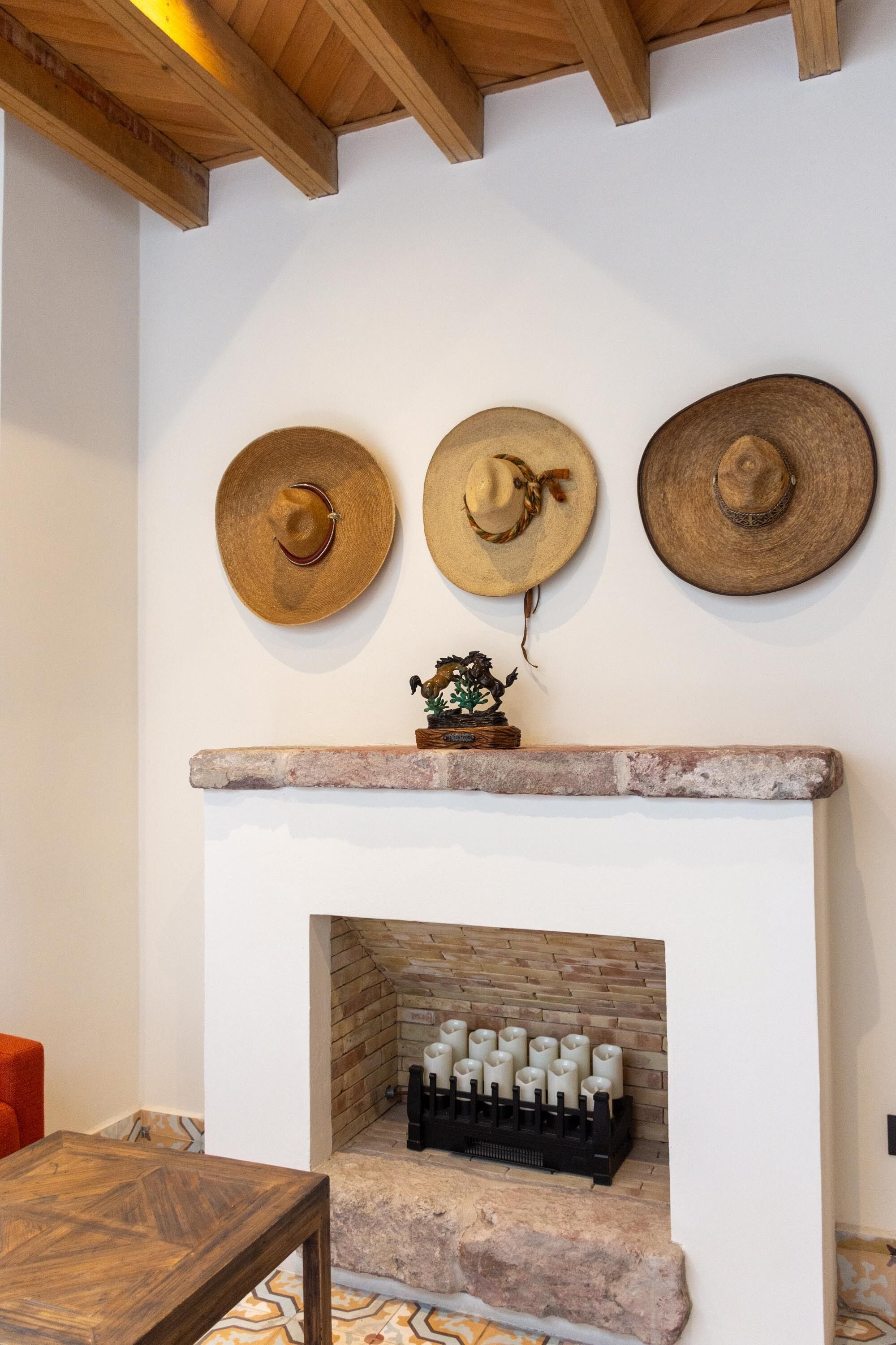 A fireplace with a stone mantle, three hats on the wall above, and a small decorative piece on the mantle.
