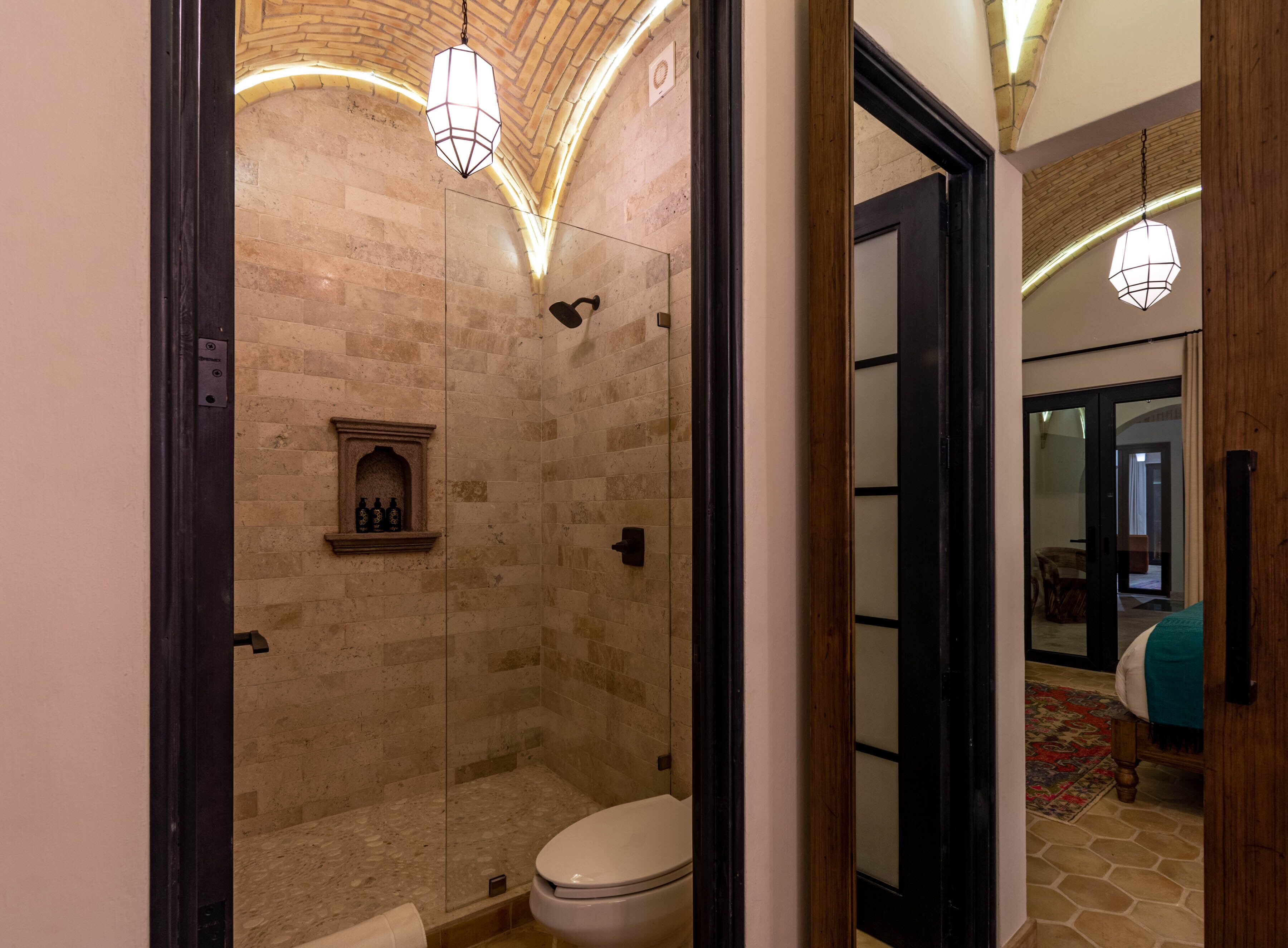 Une salle de bain moderne avec une douche à parois de verre, une étagère intégrée où sont rangées des bouteilles, et une porte en bois donnant sur une autre pièce.
