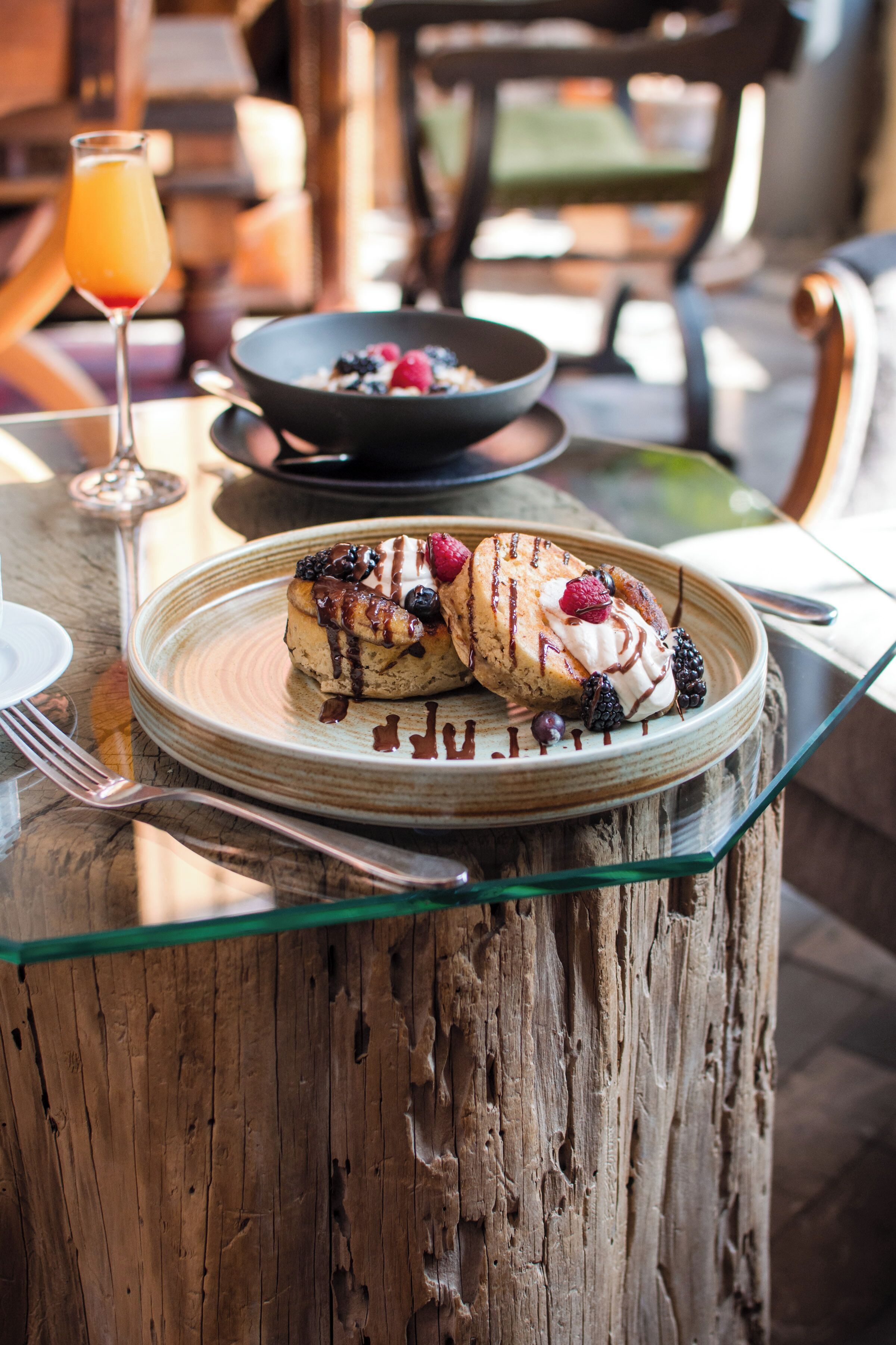 A breakfast setting with a plate of pancakes topped with berries and chocolate sauce, a glass of orange juice, and a bowl of cereal.