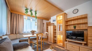 Ein gemütliches Wohnzimmer mit einer Couch, Sesseln, einem Fernseher, Holzregalen und einem Essbereich mit Tisch und Stühlen.