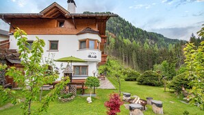 Ein zweistöckiges Holzhaus mit Balkon, umgeben von einem gepflegten Garten mit unterschiedlichen Pflanzen und einem Steinweg, der zum Eingang führt.
