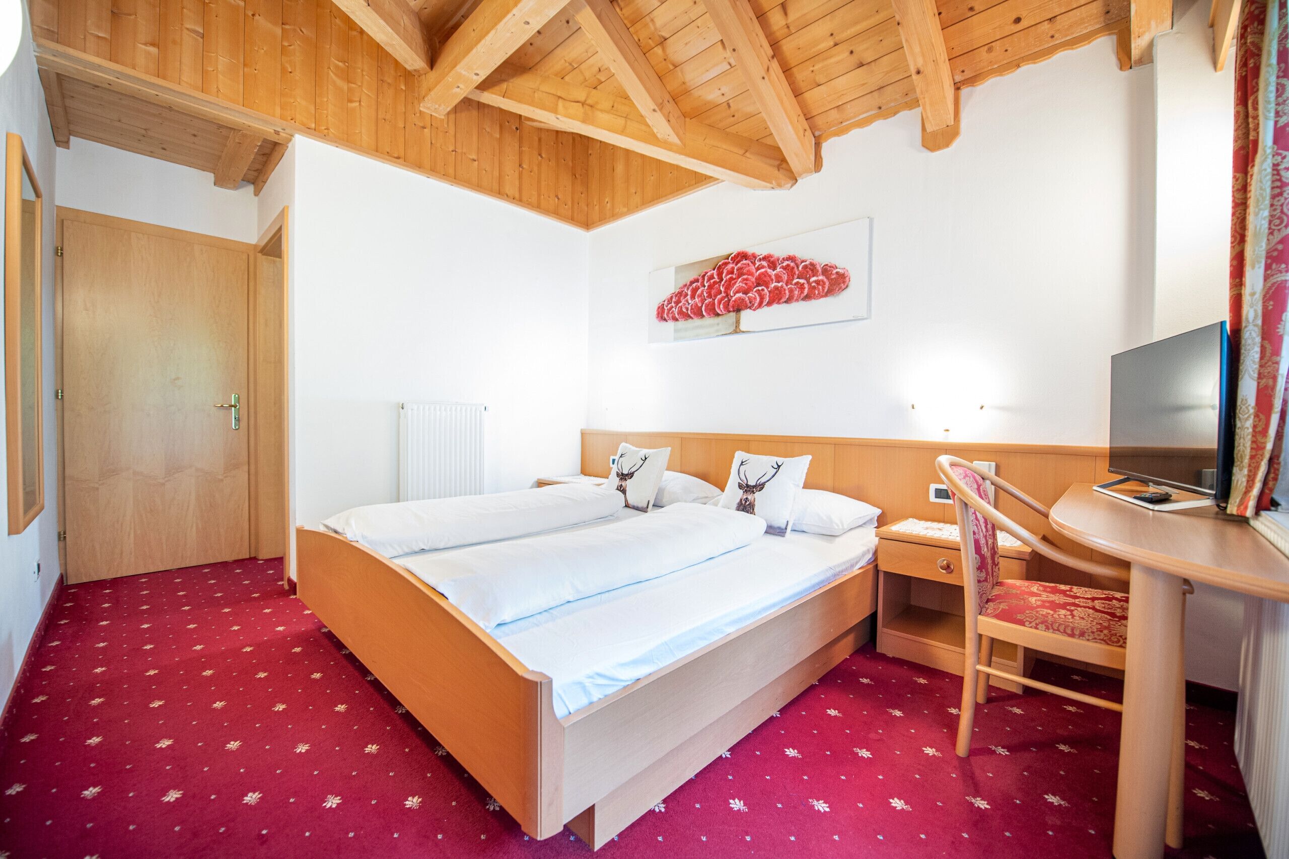 A hotel room with a wooden ceiling, a bed with white bedding, a red carpet, a small bedside table, a chair, and a television.