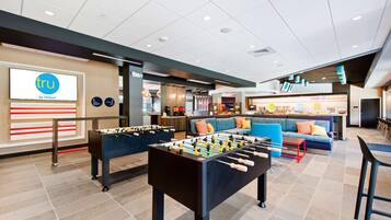 A hotel lobby with a foosball table, a seating area with sofas, and a kitchenette in the background.