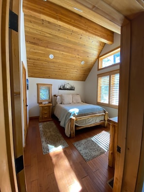 A bedroom with a wooden ceiling, a bed, a nightstand, a dresser, and a window with blinds.
