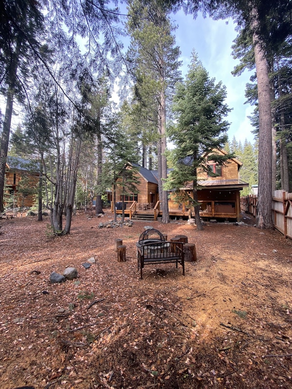 A cabin surrounded by tall pine trees, with a wooden deck and a fire pit in the foreground.