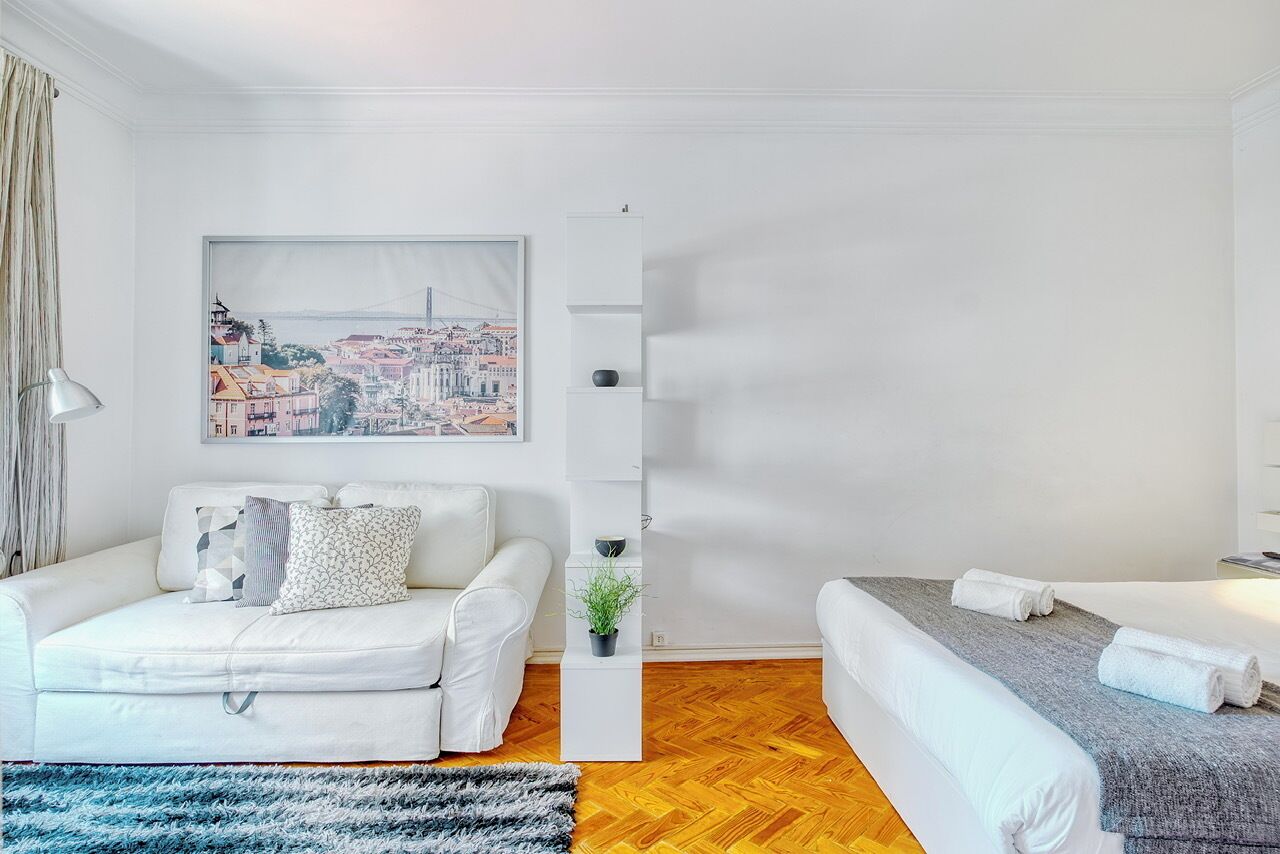 Een moderne hotelkamer met een witte bank, een bed met een grijze deken, een stadslandschap schilderij aan de muur, en een kleine plant op een witte bijzettafel.