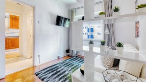 Ein modernes Apartment mit Küche, einem an der Wand montierten Fernseher, einem Balkon mit Topfpflanzen und einer Couch mit Kissen.