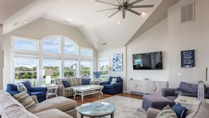 A spacious living room with a high ceiling, large windows, and a television. - Destination-OBX Soundfront, 8 bdrm, 10 bth, elavator, pool, hot tub, game room (Nags Head)